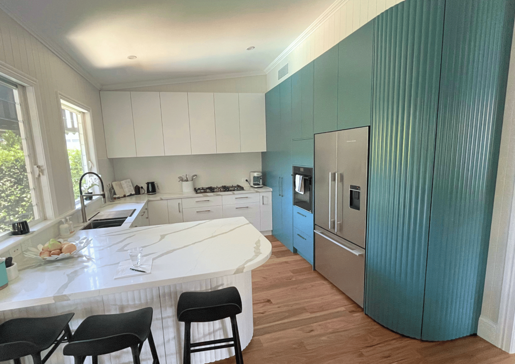 Modern kitchen renovation with white and teal cabinetry, stainless steel appliances, marble island countertop, and black stools on wooden flooring.