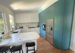 Modern kitchen renovation with white and teal cabinetry, stainless steel appliances, marble island countertop, and black stools on wooden flooring.